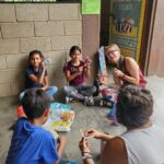 Woman and children making yarn crafts sit in circle smiling at camera